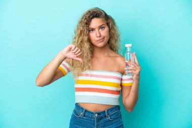 Mavi arka planda izole edilmiş su şişesi olan genç sarışın kadın olumsuz bir ifadeyle baş parmağını gösteriyor.