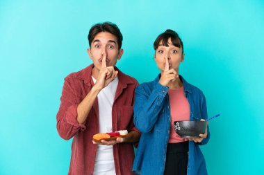 Mavi arka planda saşimi ve ramen tutan genç melez arkadaşlar sessiz bir jest işareti gösteriyorlar. Parmağını ağzına sok.