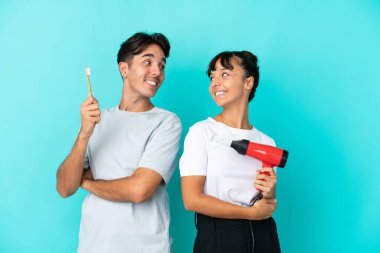 Mavi arka planda izole edilmiş saç kurutma makinesi ve diş fırçası tutan genç ve melez bir çift, gülümseyerek omuzlarının üzerinden bakıyor.