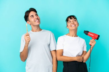 Mavi arka planda saç kurutma makinesi ve diş fırçası tutan genç bir çift gülümserken yukarı bakıyor.