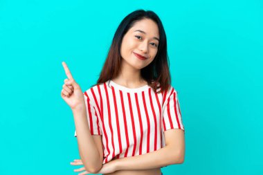 Genç Vietnamlı kadın mavi arka planda izole edilmiş ve parmağını kaldırıyor.
