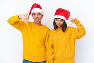 Genç melez çift, beyaz arka planda izole edilmiş bir şekilde Noel 'i kutluyor. İki eliyle baş parmağını aşağı gösteriyor.
