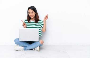 Yerde dizüstü bilgisayarı olan genç Vietnamlı bir kadın beyaz duvarda izole bir şekilde oturuyor. Yan tarafları işaret ediyor ve mutlu.
