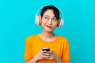 Genç Vietnamlı kadın mavi arka planda izole edilmiş. Cep telefonuyla müzik dinliyor ve düşünüyor.