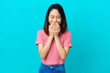 Genç Vietnamlı bir kadın mavi arka planda izole edilmiş mutlu ve gülümseyen elleriyle ağzını kapatıyor.