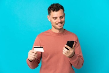 Kafkasyalı yakışıklı genç adam mavi arka planda izole edilmiş ve kredi kartıyla cep telefonuyla alışveriş yapıyor.