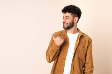Bej arka planda izole edilmiş genç Faslı bir adam bir ürün sunmak için kenarı işaret ediyor.