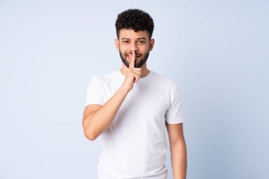 Faslı genç bir adam mavi arka planda izole edilmiş. Parmağını ağzına koyduğunda sessizlik işareti gösteriyor.