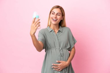 Genç Uruguaylı kadın mavi arka planda izole edilmiş hamile ve elinde şaşırtıcı bir ifadeyle bir beslenme şişesi tutuyor.