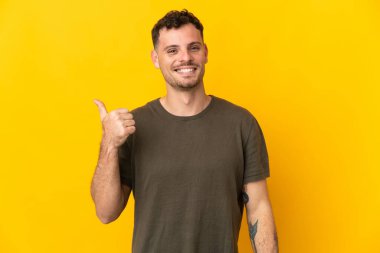 Sarı arka planda izole edilmiş beyaz, yakışıklı genç bir adam bir ürün sunmak için kenarı işaret ediyor.