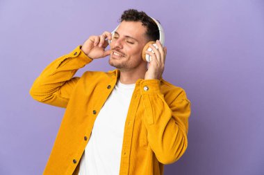 Genç, beyaz, yakışıklı bir adam mor arka planda izole edilmiş müzik dinliyor ve şarkı söylüyor.