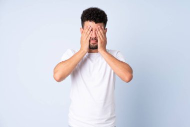 Faslı genç bir adam mavi arka planda izole edilmiş. Gözlerini elleriyle kapatıyor.
