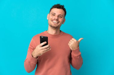Genç, beyaz, yakışıklı bir adam mavi arka planda izole edilmiş, cep telefonu kullanıyor ve yan tarafı işaret ediyor.