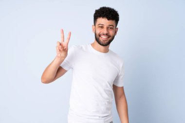 Mavi arka planda izole edilmiş genç Faslı adam gülümsüyor ve zafer işareti gösteriyor.