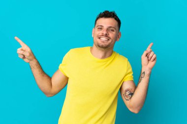 Mavi arka planda izole edilmiş beyaz tenli yakışıklı adam parmak uçlarını işaret ediyor ve mutlu.