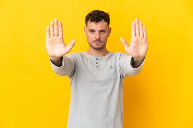 Sarı arka planda izole edilmiş beyaz, yakışıklı genç adam dur hareketi yapıyor ve hayal kırıklığına uğruyor.