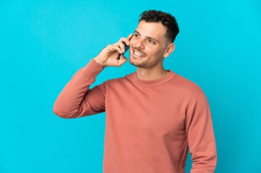 Beyaz tenli yakışıklı adam mavi arka planda izole edilmiş cep telefonuyla biriyle sohbet ediyor.