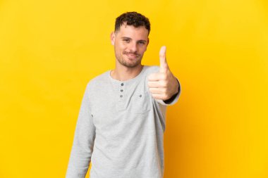 Beyaz tenli yakışıklı adam sarı arka planda izole edildi. Başparmakları havada, çünkü iyi bir şey oldu.