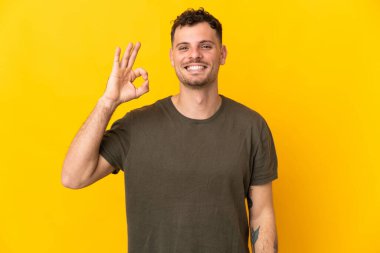 Sarı arka planda izole edilmiş beyaz, yakışıklı genç bir adam parmaklarıyla tamam işareti gösteriyor.
