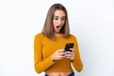 Beyaz arka planda izole edilmiş genç beyaz bir kadın kameraya bakarken şaşırmış bir ifadeyle cep telefonunu kullanıyor.