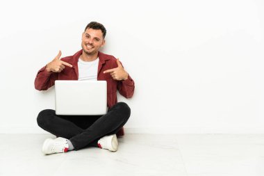 Genç, yakışıklı, beyaz bir adam dizüstü bilgisayarlı, gururlu ve kendini beğenmiş bir şekilde yerde oturuyor.