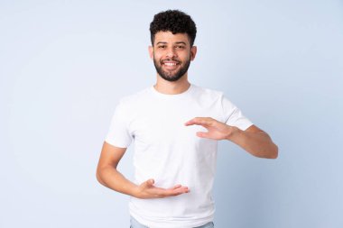 Faslı genç bir adam mavi arka planda izole edilmiş. Avucunda hayali bir şey tutuyor. Bir ilan vermek için.