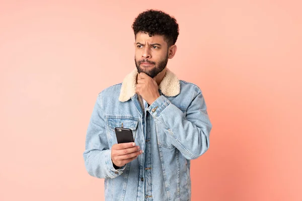疑いと思考を持つピンクの背景に隔離された携帯電話を使用して若いモロッコ人 — ストック写真