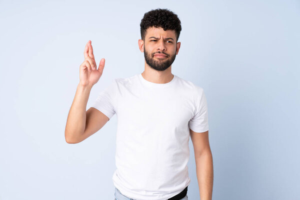 Молодой марокканский человек изолирован на синем фоне с скрещиванием пальцев и желанием лучшего