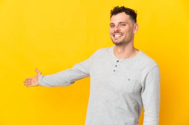 Sarı arka planda izole edilmiş beyaz tenli yakışıklı adam, davet edilmek için ellerini yana uzatıyor.