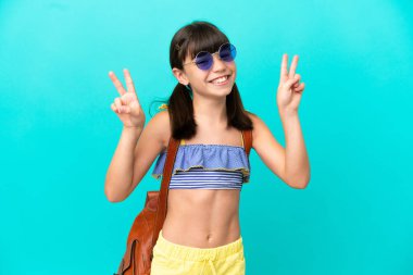 Küçük beyaz bir çocuk kumsala gidiyor. Mavi arka planda tek başına. İki eliyle zafer işareti gösteriyor.