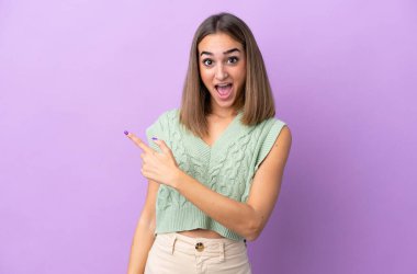 Mor arka planda izole edilmiş genç beyaz kadın şaşırmış ve işaret ediyor.