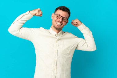 Mavi arka planda izole edilmiş beyaz, yakışıklı genç bir adam güçlü bir jest yapıyor.
