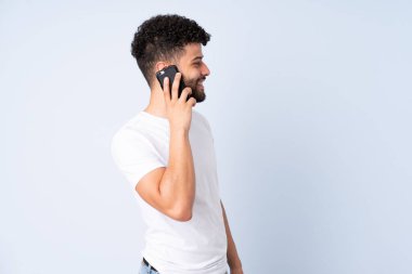 Faslı genç bir adam mavi arka planda izole edilmiş ve cep telefonuyla sohbet ediyor.