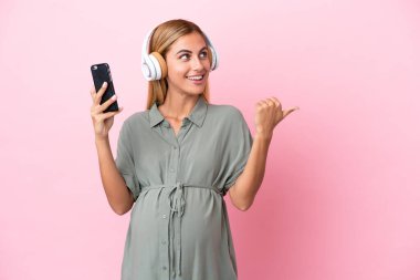 Genç Uruguaylı kadın mavi arka planda izole edilmiş hamile ve işaret ederken müzik dinliyor.