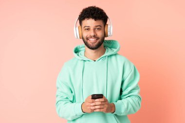 Genç Faslı bir adam pembe arka planda izole edilmiş. Cep telefonuyla müzik dinliyor ve ön tarafa bakıyor.