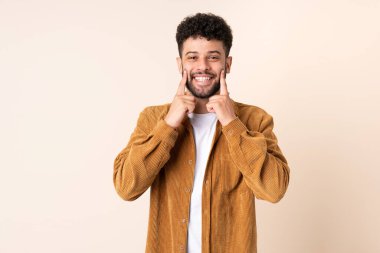 Bej arka planda izole edilmiş genç Faslı adam mutlu ve hoş bir ifadeyle gülümsüyor.