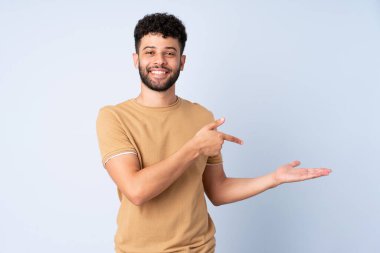 Faslı genç bir adam mavi arka planda izole edilmiş. Avucunda hayali bir şey tutuyor. Bir ilan vermek için.