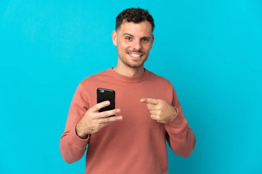 Genç, beyaz, yakışıklı bir adam mavi arka planda izole edilmiş ve cep telefonu kullanıyor.