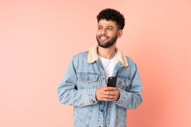 Pembe arka planda izole edilmiş cep telefonu kullanan genç Faslı adam yukarı bakarken bir fikir düşünüyor.