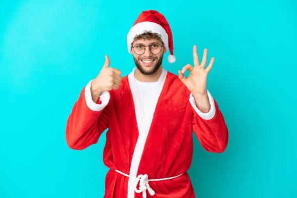 装扮成圣诞老人的年轻人在蓝色的背景上被隔离 露出可怜兮兮的手势和竖起大拇指的姿势 — 图库照片