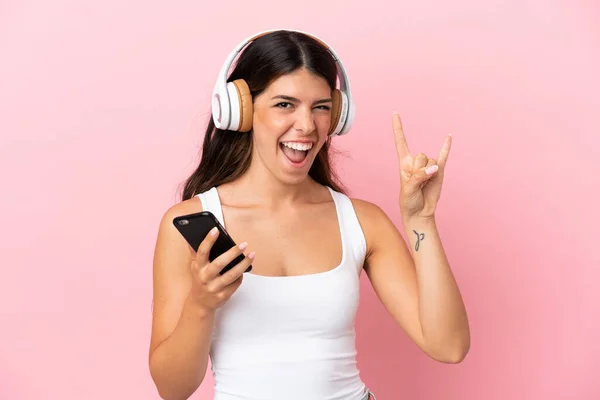 若いですCaucasian女性隔離オンピンクバックグランドリスニング音楽とともにモバイルメイキングロックジェスチャー — ストック写真