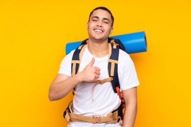 Sarı arka planda izole edilmiş büyük sırt çantalı Asyalı genç bir adam baş parmak işareti yapıyor.