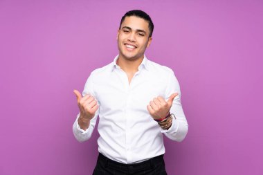 Asyalı yakışıklı adam mavi arka planda izole edilmiş, başparmakları havada ve gülümsüyor.