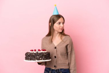 Elinde doğum günü pastası tutan Litvanyalı genç bir kadın pembe arka planda izole edilmiş bir şekilde yan tarafa bakıyor.