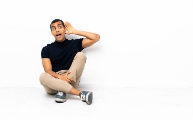 Afrika kökenli Amerikalı bir adam yerde oturup bir şeyler dinlerken elini kulağına koydu.