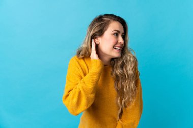 Genç Brezilyalı bir kadın mavi arka planda izole edilmiş bir şekilde elini kulağına koyarak bir şeyler dinliyor.