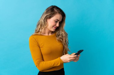 Mavi arka planda izole edilmiş genç Brezilyalı kadın cep telefonuyla mesaj gönderiyor.