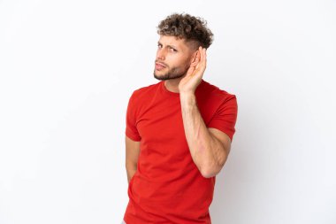 Beyaz arka planda izole edilmiş beyaz tenli yakışıklı bir adam elini kulağına koyarak bir şeyler dinliyor.