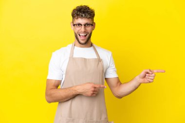 Restoran garsonu. Sarı arka planda izole edilmiş sarışın adam. Şaşırmış ve işaret ediyor.