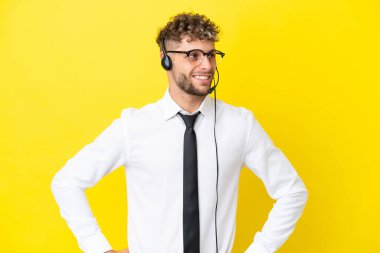 Sarı arka planda izole edilmiş, kolları kalçasında ve gülümseyen bir kulaklıkla çalışan pazarlamacı sarışın adam.